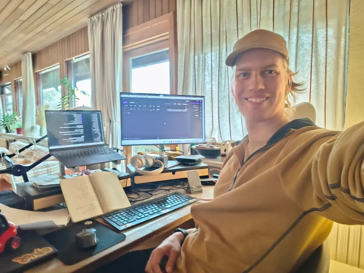 Mads sidder ved sit skrivebord med en bærbar computer, en stor skærm og en opslået notesbog og smiler til kameraet.