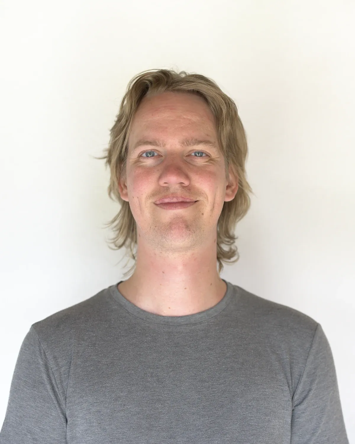 Mads Holst Jensen med halvlangt lyst hår og grå t-shirt smiler til kameraet foran en hvid væg.